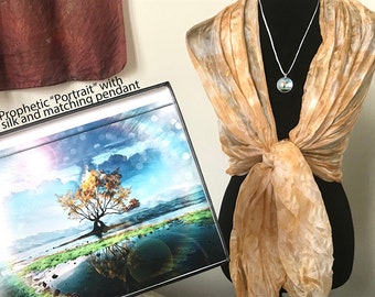 May include: A silk scarf in shades of beige and brown with a matching pendant featuring a nature scene. The scarf is tied around a mannequin's neck. The pendant is on a silver chain. The scarf and pendant are displayed with a framed print of a tree reflected in a body of water.