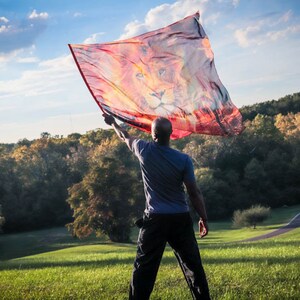 May include: A person holds a large, colorful flag with a lion's face printed on it. The flag is waving in the wind, and the person is standing in a grassy field with trees in the background.