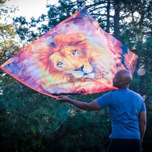 May include: A person holds a colorful fabric flag with a lion's face printed on it. The flag is flowing in the wind, and the person is standing in a forest.