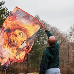 May include: A person holds a flag with a lion's face in flames. The flag is orange, yellow, and red. The person is wearing a green hoodie and grey pants. The background is a blurry forest.