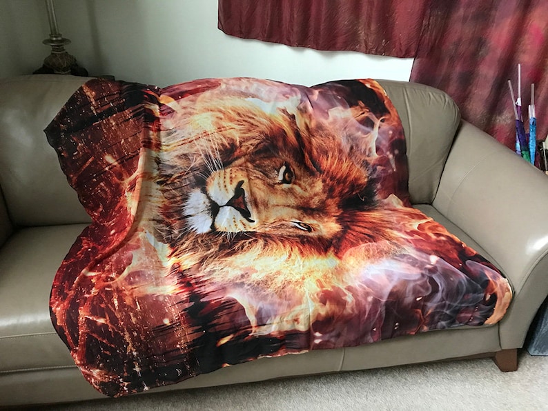 May include: A throw blanket with a fiery orange and yellow lion face print. The lion's eyes are open and the background is a swirling mix of orange, yellow, and white.