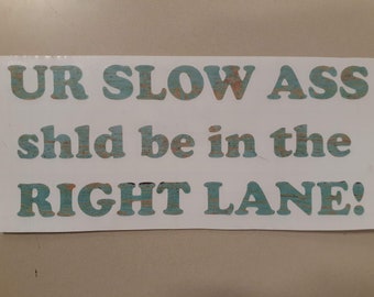 May include: A white sign with blue and orange text that reads "UR SLOW ASS shld be in the RIGHT LANE!"