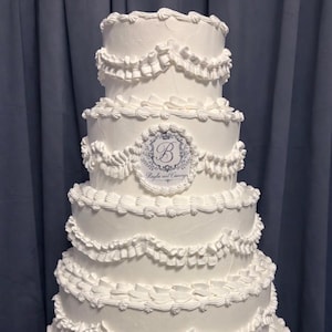 May include: A multi-tiered white cake with intricate frosting details. The cake features layers of smooth frosting, ruffled edges, and decorative swirls. A circular emblem with the letter "B" is centered on one layer. The cake is a classic design for celebrations.