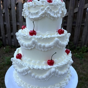 May include: A three-tiered white cake with decorative frosting and red cherries. The cake has ruffled frosting edges and is displayed on a white cake board. The cherries have green stems.