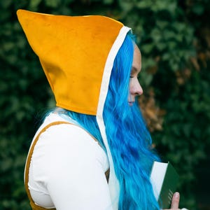 May include: A person with bright blue hair is wearing a yellow hooded garment with a white trim. They are holding a book in their hands.
