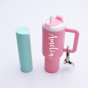 May include: A pink and white keychain shaped like a cup with a straw and a handle. The cup has the name "Amelia" written on it. Next to the cup is a light blue lip balm with the word "eos" written on it.
