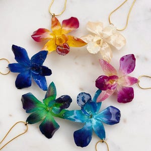 Może przedstawiać: Sześć zawieszek z kwiatów orchidei na złotych łańcuszkach. Orchidee są konserwowane i dostępne w różnych kolorach, w tym żółtym, czerwonym, białym, niebieskim, fioletowym i zielonym.