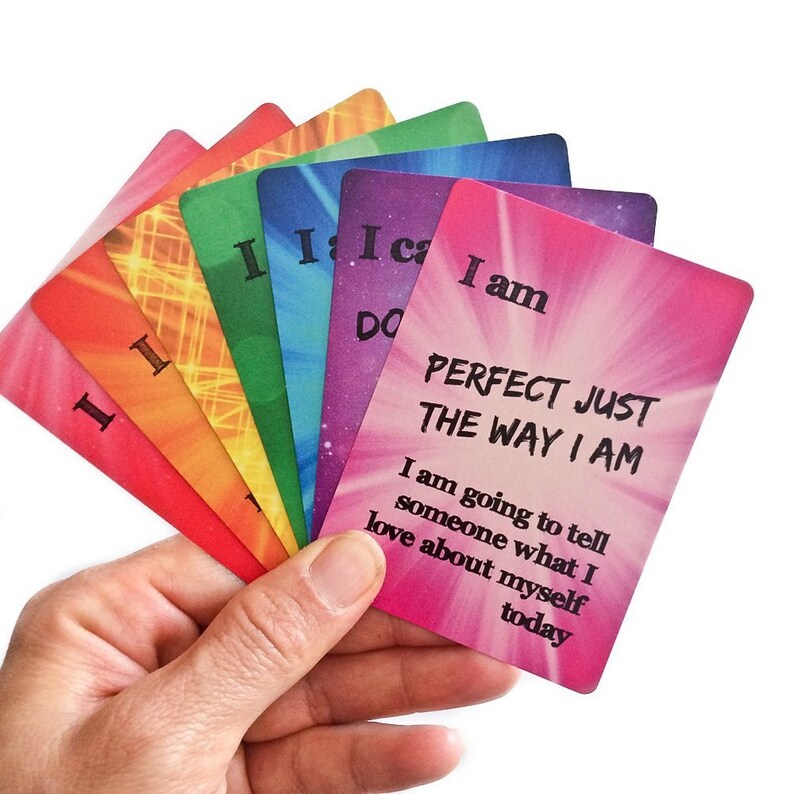 K&ouml;nnte beinhalten: Ein Satz Affirmationskarten in Regenbogenfarben mit dem Text "Ich bin perfekt, so wie ich bin". Die Karten liegen in einer Hand, aufgef&auml;chert, um die verschiedenen Farben und Botschaften zu zeigen.