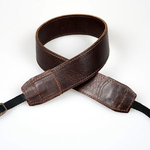 Può includere: Un cinturino per fotocamera in pelle marrone con cinturini regolabili neri e fibbie in ottone.
