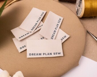 May include: A collection of rectangular, off-white fabric clothing labels featuring the black printed text DREAM PLAN SEW. These small, woven tags are stacked together, showcasing a clean, simple design intended for sewing projects and garment branding.