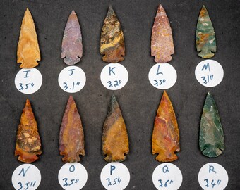 May include: A collection of 10 arrowheads made of different types of stone, each with a unique pattern and color. The arrowheads are arranged in two rows of five, with a white circle label beneath each one. The labels include letters and measurements in inches.