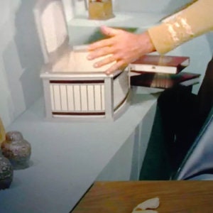 Könnte beinhalten: Eine silber-weiße Truhe mit geschwungener Front und vertikalen Lamellen. Die Hand einer Person liegt auf dem offenen Deckel. Zwei dekorative Vasen befinden sich im Vordergrund. Ein Stapel Bücher ist sichtbar.