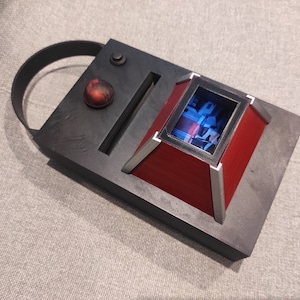 Op de afbeelding: Een futuristisch ogend apparaat met een donkergrijze rechthoekige basis en een rood, schuin display. Het heeft een zwarte handgreep, een rode bol en een zilver omlijst scherm met een blauw en wit beeld. Het apparaat is waarschijnlijk een rekwisiet of model.