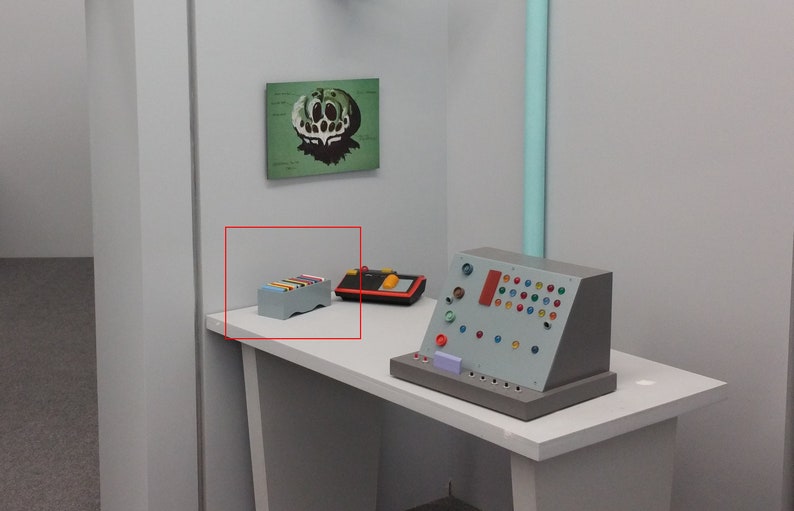 May include: A gray rectangular box with colorful rectangular blocks on top. The box is on a white table in a room with a retro-futuristic aesthetic. Other objects on the table include a control panel and a black and red device.
