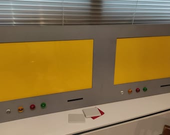 Puede incluir: Un panel gris rectangular grande con dos pantallas amarillas. El panel tiene una serie de luces y botones de colores a lo largo del borde inferior. Dos objetos cuadrados pequeños y un paño rojo están frente al panel.