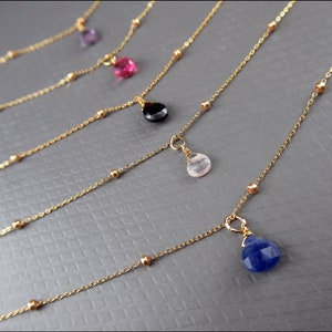 May include: Five gold necklaces with different colored gemstone pendants. The necklaces feature a delicate chain with small gold beads. The gemstones are pink, red, black, light pink, and blue.
