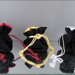 Può includere: Tre sacchetti in velluto nero con bordi in raso di diversi colori. I sacchetti sono allineati su una superficie riflettente. Il sacchetto a sinistra ha un bordo bordeaux, il sacchetto al centro ha un bordo dorato e il sacchetto a destra ha un bordo argentato.