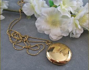 May include: A gold locket necklace with a round, shiny pendant. The necklace is made of a delicate chain and is lying on a gray surface.