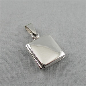 Könnte beinhalten: Ein polierter Silberanhänger in quadratischer Form mit einem Klappverschluss. Der Anhänger hat oben eine kleine Schlaufe für eine Kette. Die Oberfläche des Anhängers ist reflektierend, und das Gesamtdesign ist schlicht und elegant.