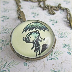 Könnte beinhalten: Eine Glaskette mit einem silbernen Kettenanhänger. Der Anhänger zeigt eine Vintage-Illustration einer Person, die einen Regenschirm hält.