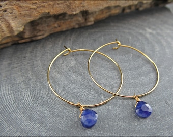 May include: Gold hoop earrings with a dangling blue gemstone. The earrings are simple and elegant.