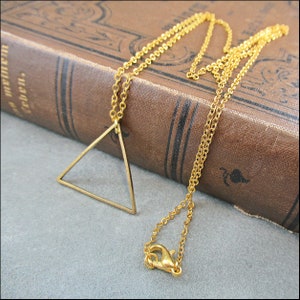 Puede incluir: Un collar de cadena dorada con un simple colgante triangular geométrico. El collar está sobre un libro encuadernado en cuero marrón.