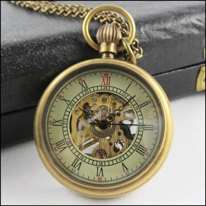 Puede incluir: Un reloj de bolsillo dorado con una esfera esqueletizada y números romanos. El reloj tiene una cadena unida a la parte superior.