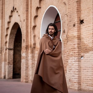 May include: A man wearing a brown hooded cloak stands in front of a brick archway. The cloak is long and flowing, and the man is looking off to the side.