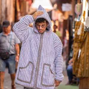 Op de afbeelding: Een persoon draagt een wit en paars gestreept hooded jack met een ritssluiting en twee zakken. Het jack heeft een bruine bies rond de zakken en de ritssluiting. De persoon draagt een bril en heeft donker haar.