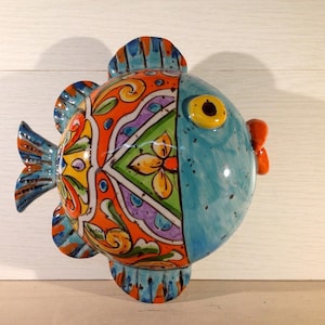 Könnte beinhalten: Eine farbenfrohe Keramik-Fisch-Wanddekoration mit blauem Körper, orangefarbenen und gelben Akzenten und weißem Hintergrund. Der Fisch hat ein großes, gelbes Auge und einen kleinen, roten Mund.