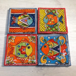 May include: Set of four colorful ceramic tiles featuring hand-painted fish designs. Each tile has a different background pattern and color scheme, including orange, blue, green, and yellow. The fish are stylized and have bright, bold colors.