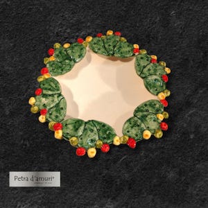 Può includere: Un piatto in ceramica con un'apertura centrale, circondato da elementi a forma di cactus verdi con dettagli rossi e gialli. Il piatto è posto su uno sfondo scuro e strutturato. Il testo "Petra d'amuri" è visibile in basso a sinistra.