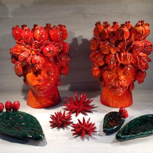May include: Two ceramic head sculptures with red floral headdresses and orange faces. Green ceramic cactus-shaped dishes and red star-shaped ornaments are arranged in front of the sculptures. The pieces have a glossy finish.