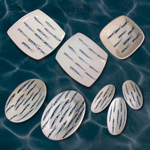 Puede incluir: Conjunto de platos y fuentes de cerámica blanca con diseño de peces pintados a mano en azul y negro. Los platos son de varias formas, incluyendo cuadrados, ovalados y oblongos. El texto "Petra d'amuri" está en la esquina superior izquierda.