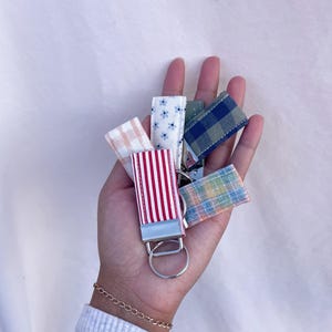 May include: A collection of fabric keychains in various patterns and colors, including red and white stripes, blue floral, and plaid designs. Each keychain has a silver metal ring.