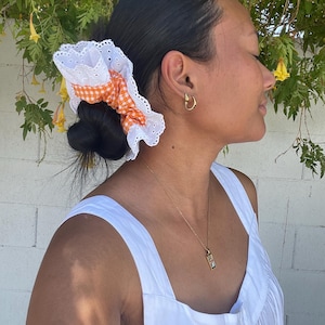 May include: A white eyelet scrunchie with orange gingham fabric and a white lace trim. The scrunchie is worn in a bun. The person is wearing a white dress and gold jewelry.