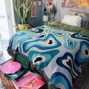May include: A woven throw blanket with a blue, teal, and beige abstract pattern is draped over a bed. A green record player and a pink vinyl record are on a wicker trunk in front of the bed. The room has art and plants.