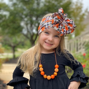 Può includere: Una bambina che indossa un vestito nero con maniche a sbuffo e una fascia per capelli a tema zucca. Indossa anche una collana di perline arancioni e dorate.