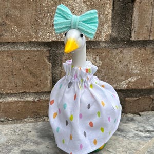 May include: A white goose figurine with a yellow beak, wearing a light mint green textured bow on its head and a white dress featuring a colorful pattern of small orange, yellow, gray, and light blue brushstroke-style spots around its neck.