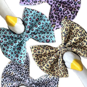 May include: Five leopard print bow ties in various colours, including purple, teal, and yellow. The bows have black and brown spots. They are arranged around white goose heads with yellow beaks.