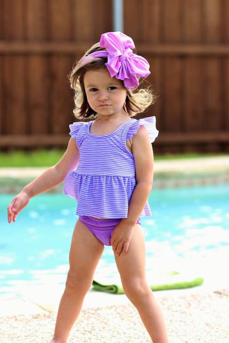 May include: A young girl wearing a purple and white striped swimsuit with a purple ruffle trim. She is wearing a large purple bow in her hair and is standing in front of a swimming pool.