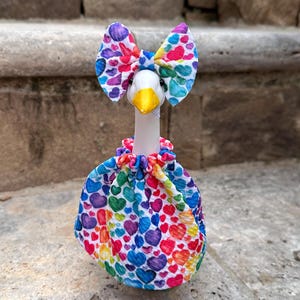 May include: A white goose figurine wearing a fabric dress and a matching bow, both featuring a vibrant, multi-colored heart pattern. The goose has a bright yellow beak and black eyes, with the dress gathered at the neck in a ruffled style.