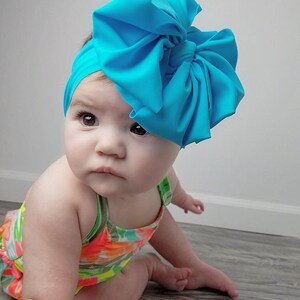 May include: A baby wearing a bright turquoise bow headband and a colorful swimsuit. The headband is made of soft fabric and has a large, voluminous bow.