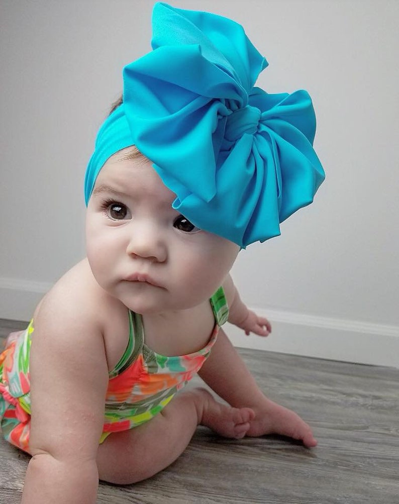 May include: A baby wearing a bright turquoise bow headband and a colorful swimsuit. The headband is made of soft fabric and has a large, voluminous bow.