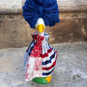 Pode incluir: Estatueta de ganso de cerâmica com um turbante de tecido azul e um vestido de retalhos com padrões vermelhos, brancos e azuis. O ganso tem um bico amarelo e uma base verde, sobre um fundo de pedra.