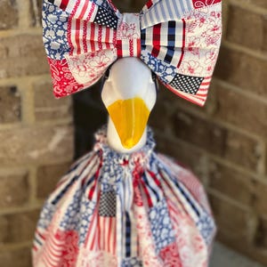 Puede incluir: Un ganso blanco con pico amarillo, con una gran pajarita y un vestido de tela estampada en rojo, blanco y azul. La pajarita y el vestido presentan diseños de bandera americana y florales.