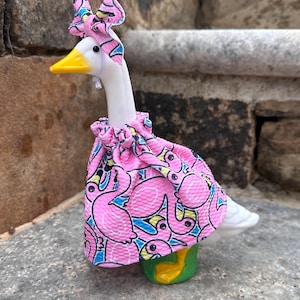 May include: A white goose figurine wearing a pink fabric outfit featuring a repeating flamingo pattern. The goose has a yellow beak and a matching pink flamingo-patterned bow tied around its head. The outfit is gathered at the neck with a ruffle.