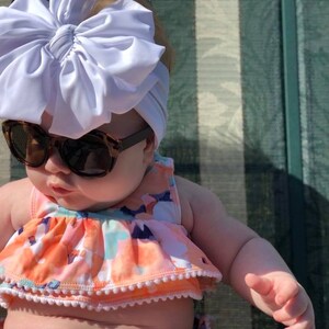 May include: A baby wearing a white bow headband and sunglasses, with a floral print swimsuit. The baby is sitting on a striped chair.