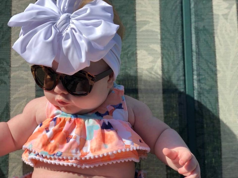 May include: A baby wearing a white bow headband and sunglasses, with a floral print swimsuit. The baby is sitting on a striped chair.
