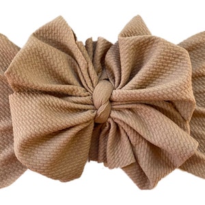May include: A large, brown, fabric bow headband. The bow is made of a textured fabric and is tied in a knot in the centre.